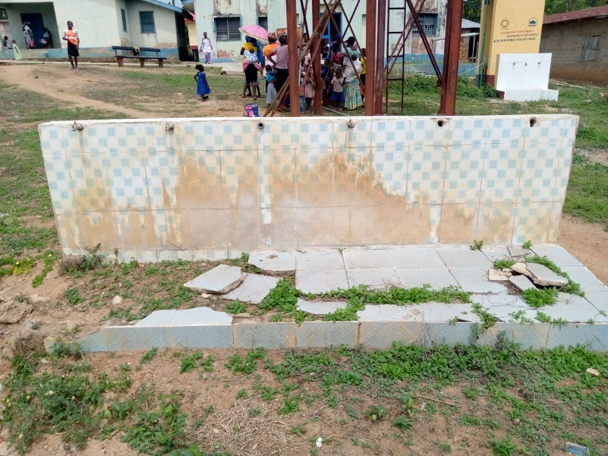 A failed public tap stand in Ijero Local Government Area, Ekiti State: six taps on a tiled stand, none of them working, with broken paving and weeds in front and the community gathered behind