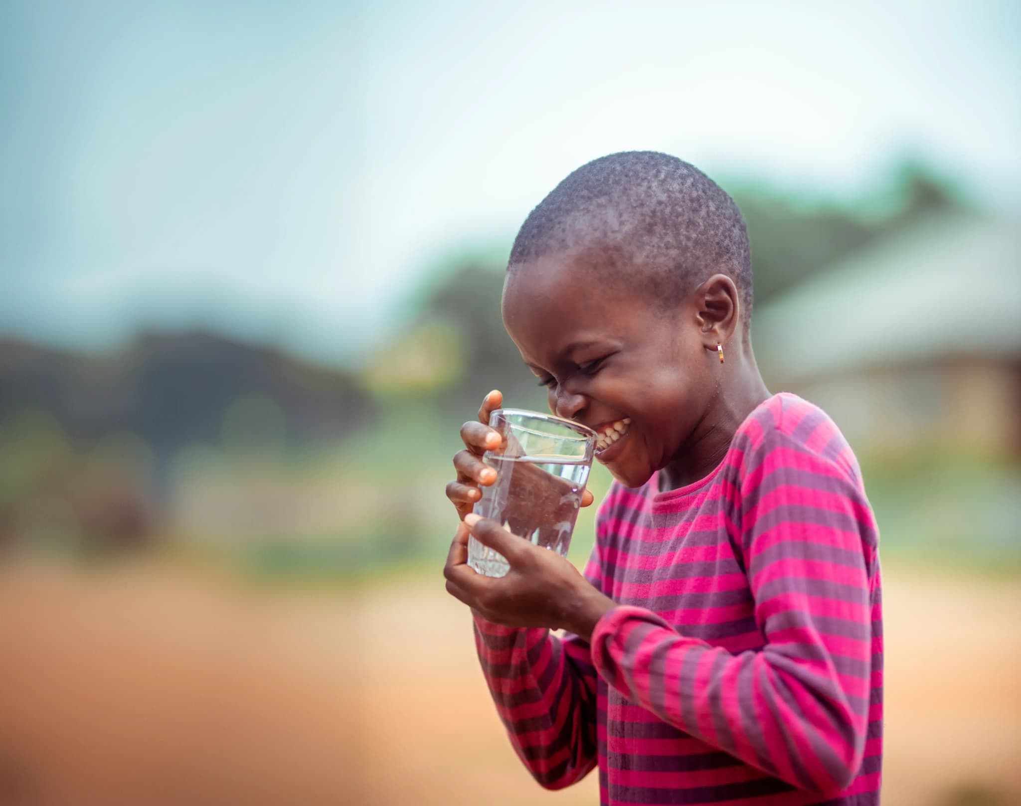 A Fairaction community member, a smiling child in a pink striped shirt, holding a glass of clean water outdoors.