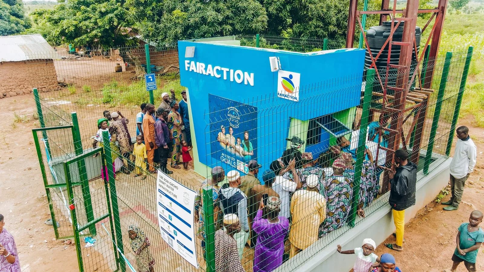 Aerial view of community gathered around the newly inaugurated Lube-Boni Igboho Smart Water Infrastructure in Oyo State, Nigeria. The Fairaction-branded kiosk stands inside a fenced compound with an elevated water tank, surrounded by community members in traditional dress.