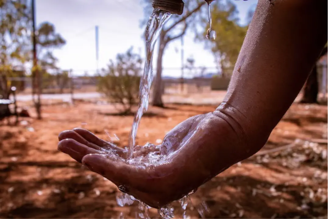 Water pours from a bottle into a hand in a dry, outdoor environment with blurred trees and soil in the background.
